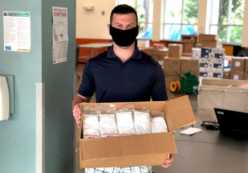 man stands holding a box of supplies wearing a black mask. Stacks of boxes are in the background.