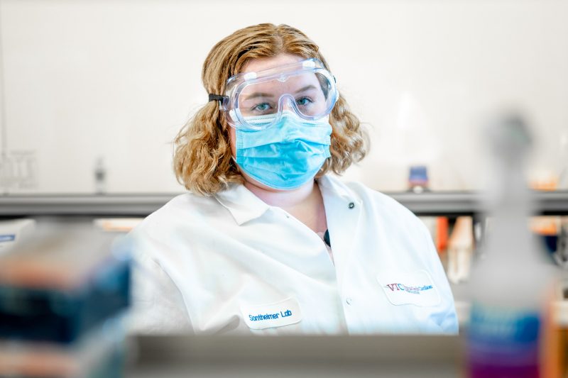 Pollack wears goggles, mask, and lab coat while standing, looking through self with lab supplies.