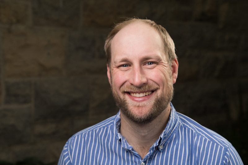 man smiles at camera with stone wall background wearing a blue striped shirt