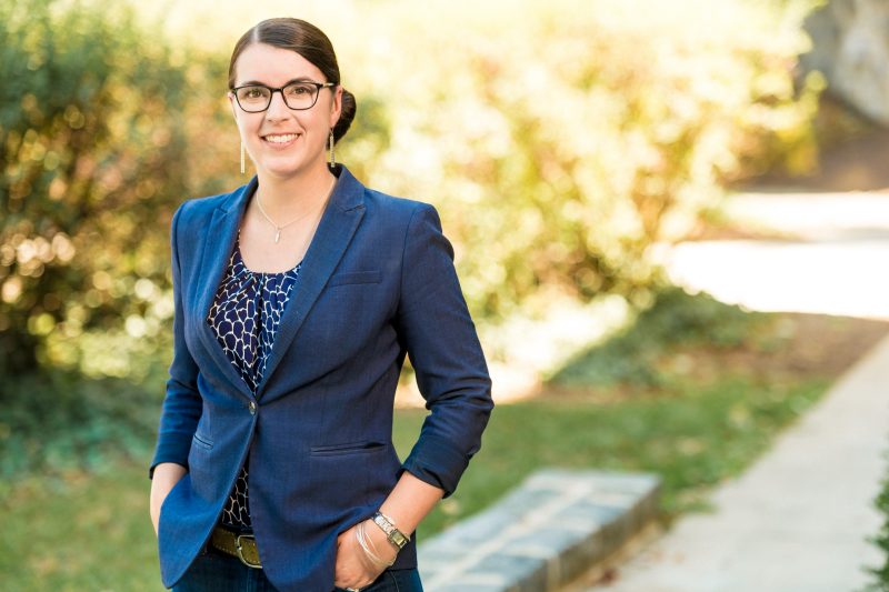 woman stands with hands in pockets and smiles, wearing glasses, blue blazer and blue shirt