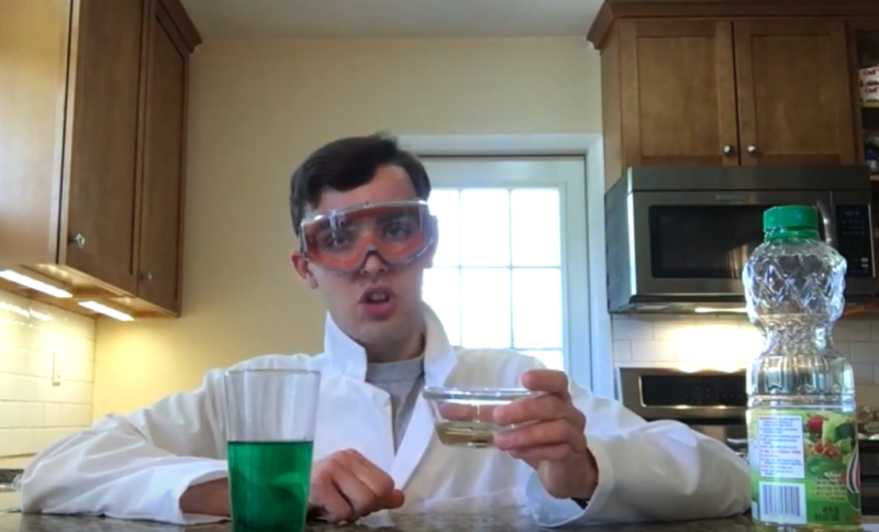Newhouse sits in kitchen with lab coat and goggles performing a chemistry demonstration
