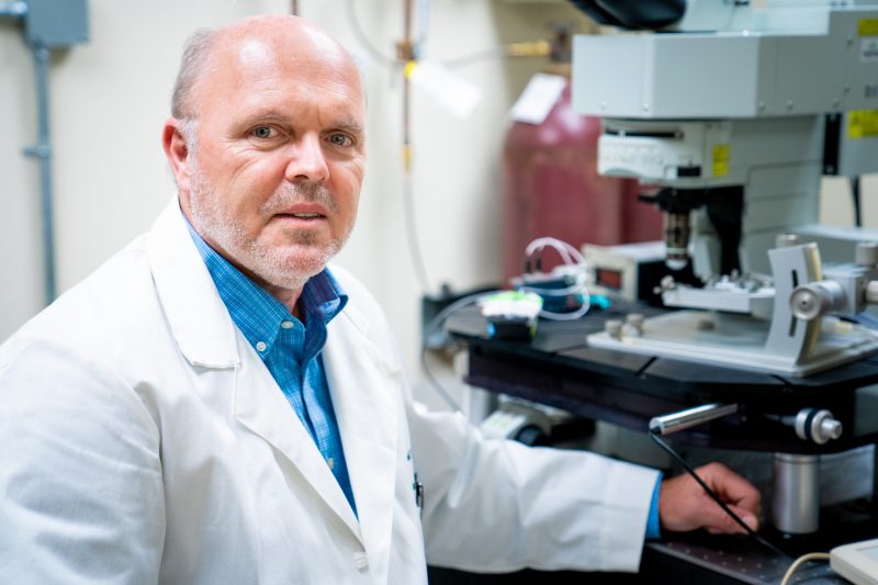 man, wearing a lab coat, sits next to microscope and turns toward camera