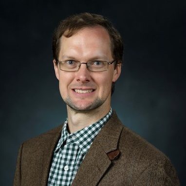 man wears glasses, tweed jacket, and plaid shirt and smiles at camera