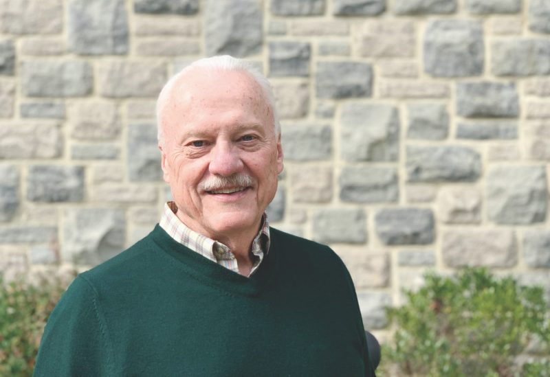 older man in dark green sweater and collared shirt outside in front of Hokie Stone wall