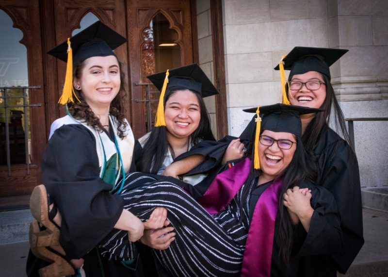 3 woman hold up their friend and smile. all wear graduation attire