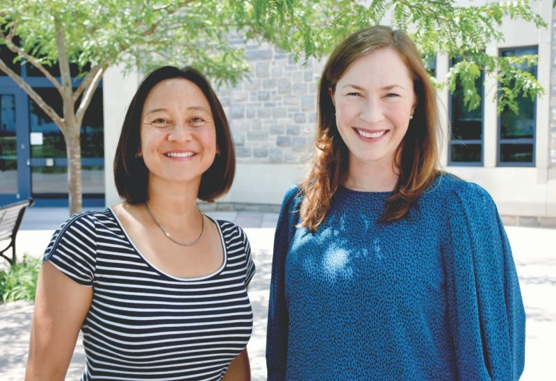 2 women stand under a tree in courtyard on sunny day and smile