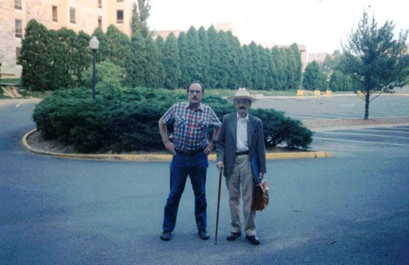 2 men in parking lot, one stands with a cane and briefcase