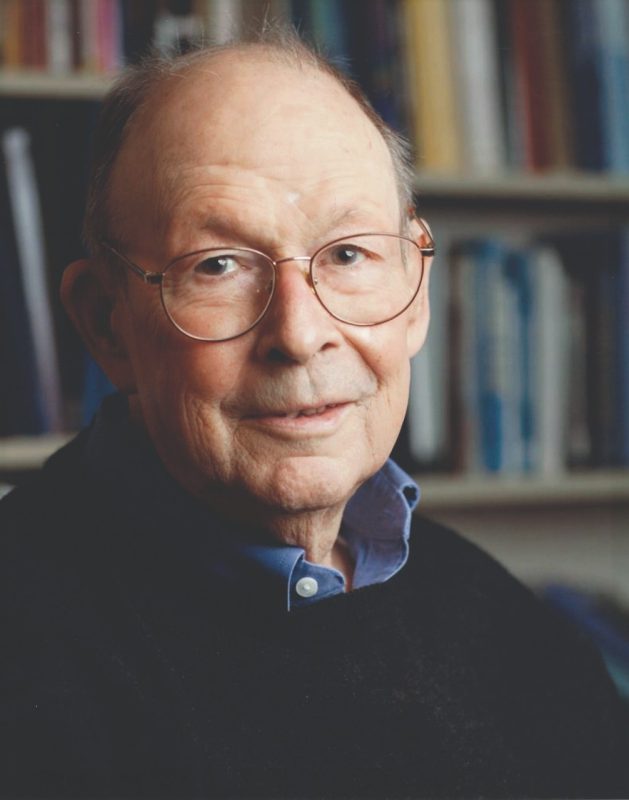 portrait of elderly man with sweater and collared shirt in front of a bookshelf