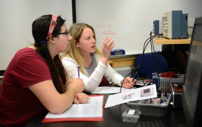 Two female students talk and look at device readout during physics lab.