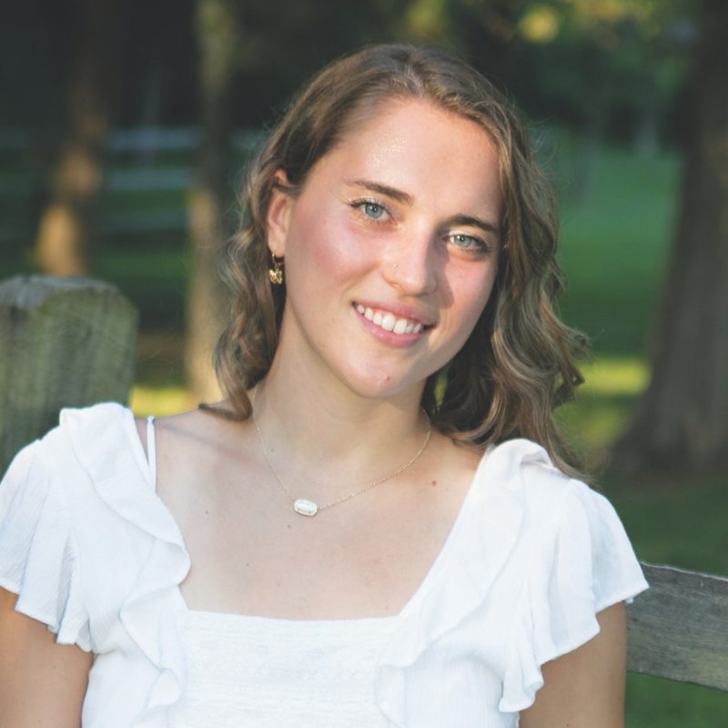 Emma stands by wood fence with trees in background in white dress.