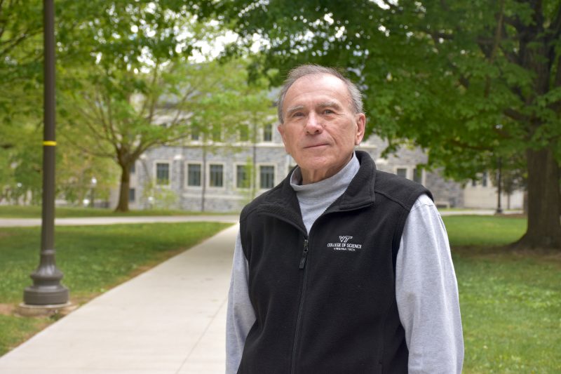 older man in fleece vest stands outside surrounded by trees on a college campus