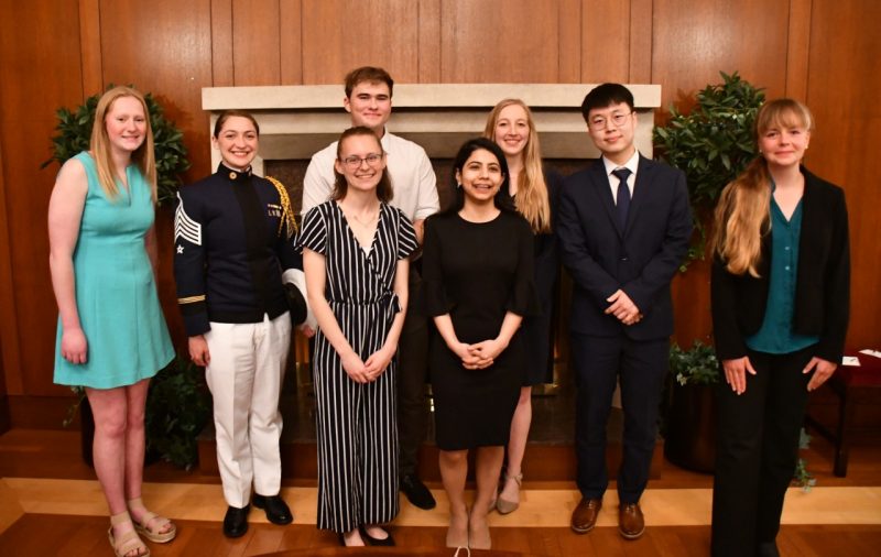 Standing in front of a fireplace at Virginia Tech Alumni Hall are , left, to right , Katie Duffett, Claire Seibel, Morgan Atkinson, Timothy Proudkii, Delshad Schroff, Janey Dike, Zhijie Yang, and Abigail Lewis. All are dressed in business attire or causal business attire, except for Sielbel who is wearing her Corps of Cadets uniform. 