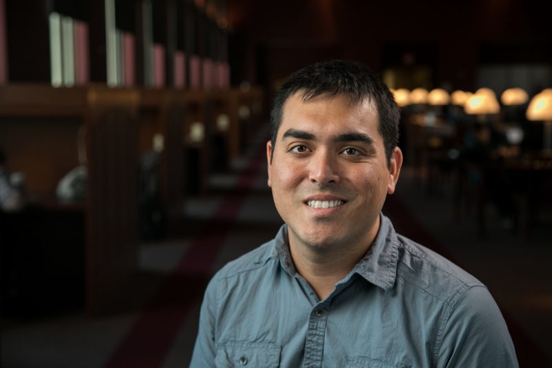 man smiles at camera with library lights and tables receding in the background